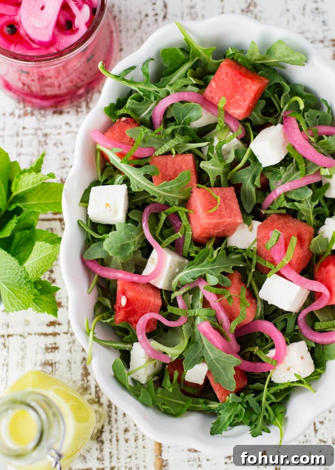 A white serving bowl with watermelon arugula salad. Feta cheese and pickled onion are in the salad as well. It's drizzled with poppyseed dressing. Mint, a bottle of dressing and pickled onions sit next to the bowl.