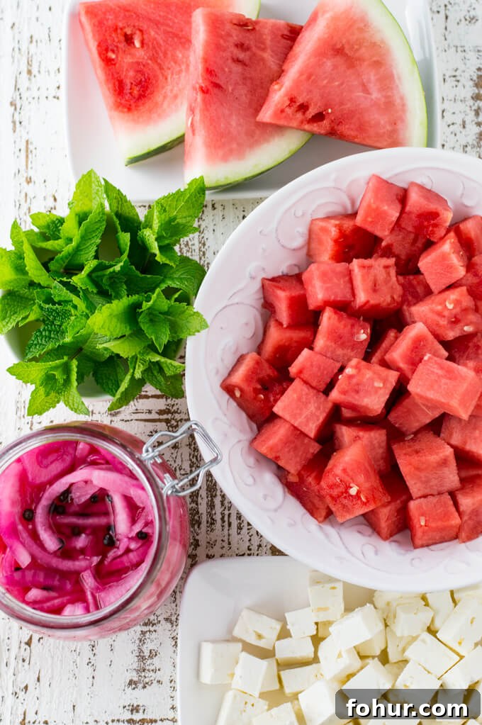A white bowl of cubed watermelon, 3 sliced watermelon pieces, fresh mint and a jar of pickled onions. Cubed feta cheese is on a small white platter.