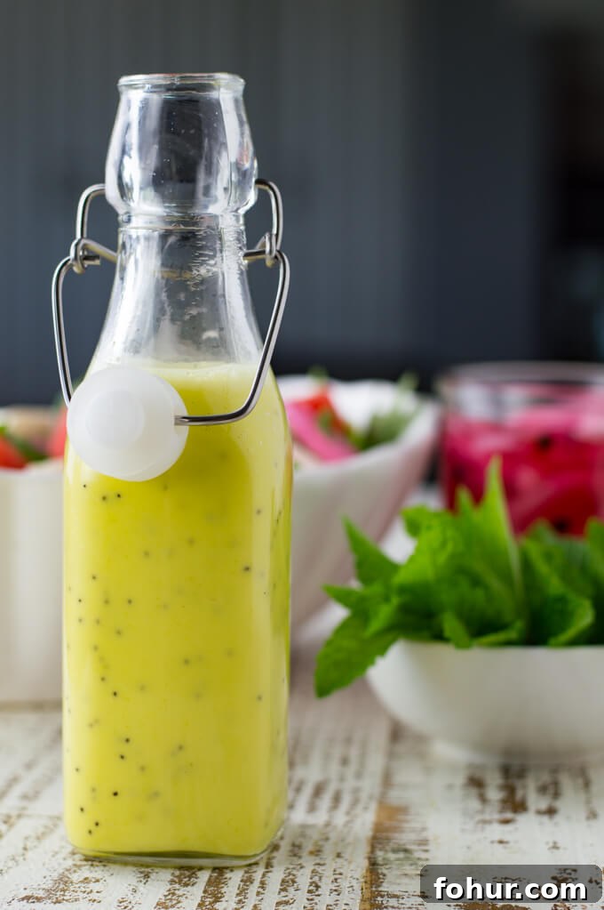 A bottle of poppyseed dressing with fresh mint, pickled onions and a watermelon salad blurred in the background.