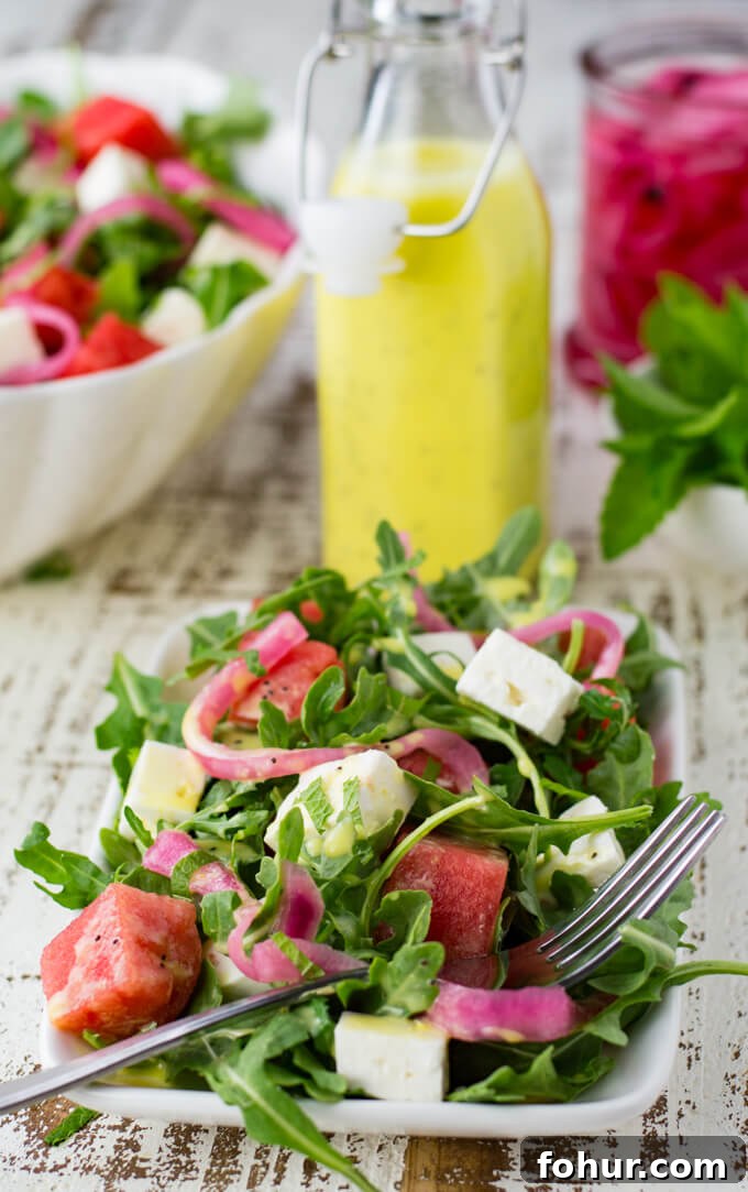 A white plate with watermelon arugula salad. Feta cheese and pickled onion are in the salad as well. It's drizzled with poppyseed dressing. Mint, a bottle of dressing and pickled onions sit next to the bowl.