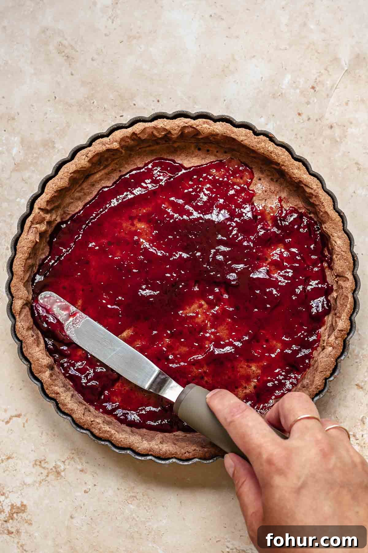 From Scratch Chocolate Raspberry Tart Recipe 6 An offset spatula spreads raspberry jam onto a cooked tart crust.