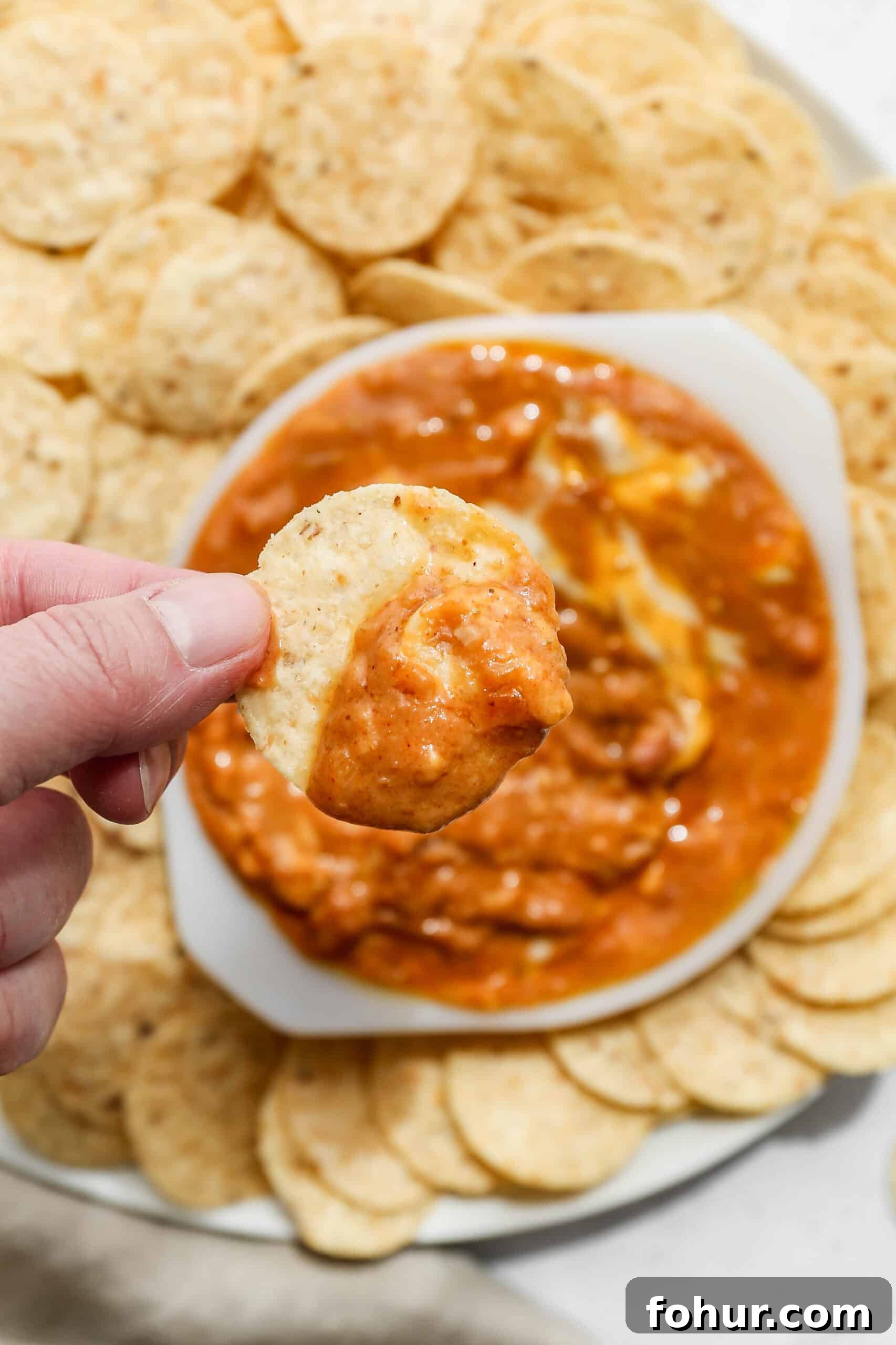 a white bowl of gluten free cheesy pinto beans surrounded by round tortilla chips with me holding a chip with dip on it.