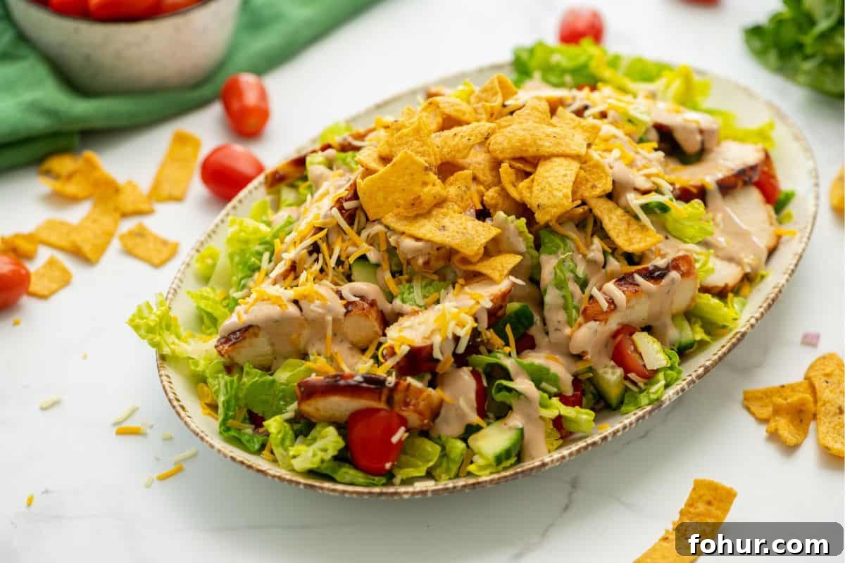Bed of lettuce topped with BBQ Chicken, shredded cheese, corn chips, and BBQ Ranch Dressing with tomatoes in background.