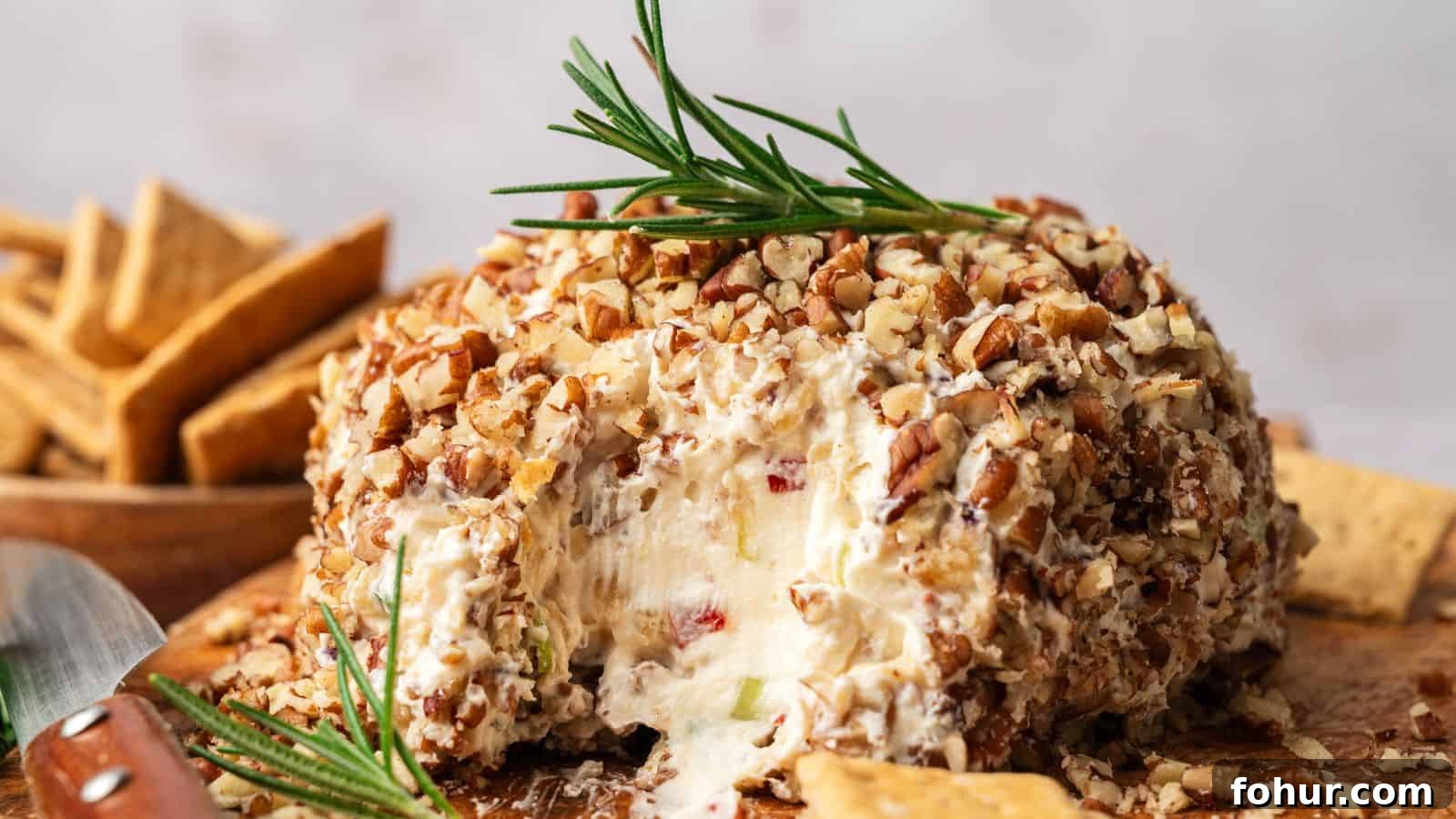31 New Year's Eve Snacks for Late-Night Parties 22 A round cheese ball coated in chopped nuts, garnished with a sprig of rosemary, sits on a wooden board with crackers beside it; a portion is scooped out.