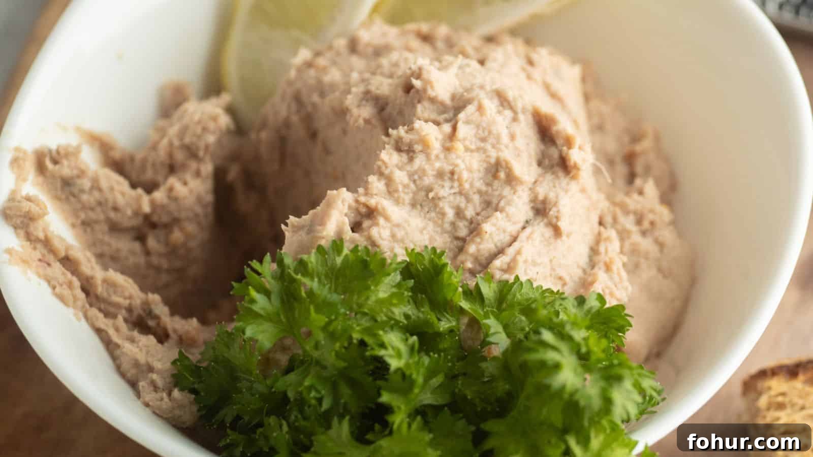 31 New Year's Eve Snacks for Late-Night Parties 4 A white bowl filled with a creamy, light brown spread, garnished with fresh parsley and lemon slices.