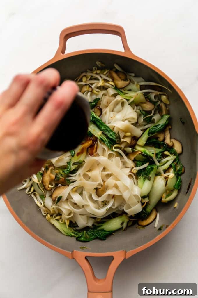 stir-fry sauce being poured into a pan of veggies and rice noodles