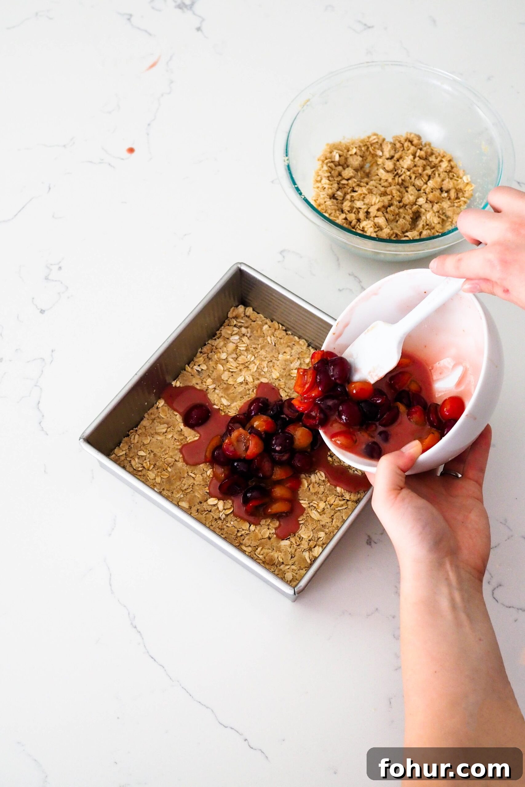 Cherry Crumble Bars with Buttery Oat Crust 9 Two hands pour cherry filling into a pan with oat crumble.