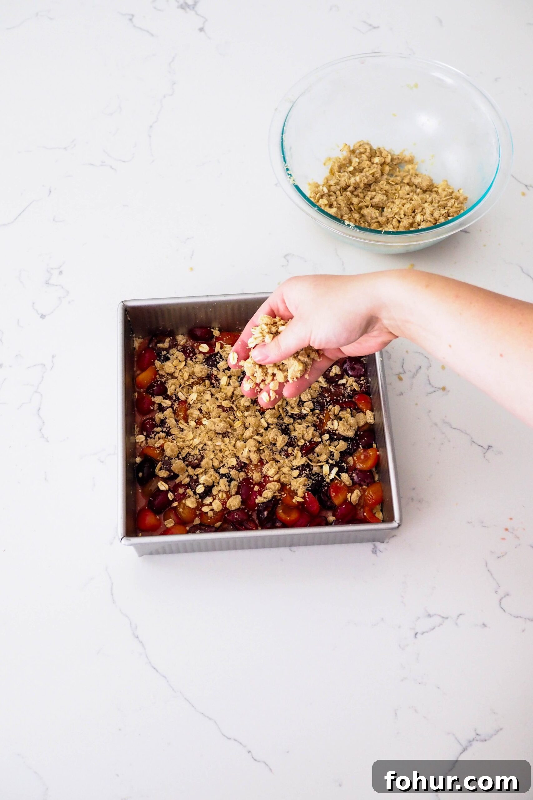 Cherry Crumble Bars with Buttery Oat Crust 10 A hand sprinkles crumble on top of a cherry filling in a pan.