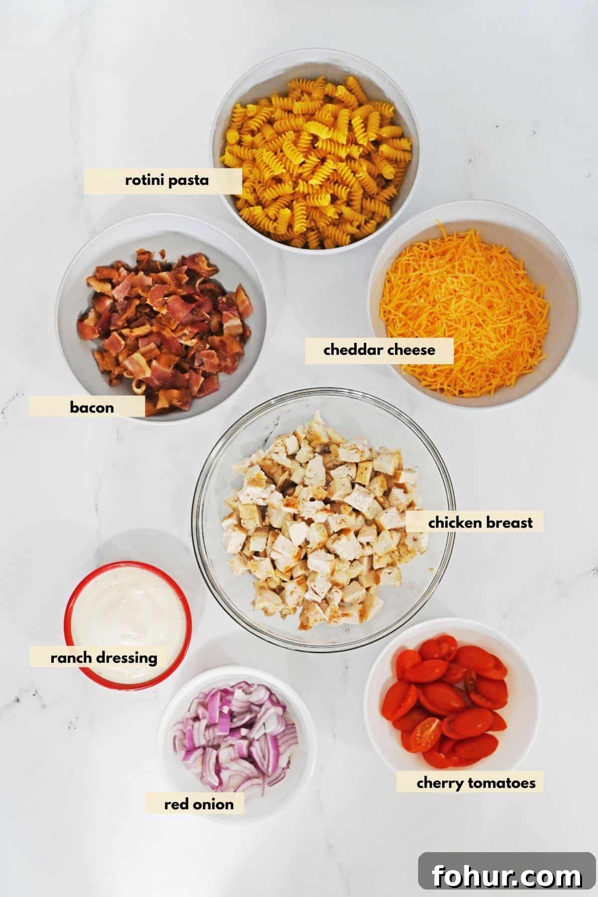 Ingredients to make pasta salad with ranch dressing, bacon, red onion, tomatoes, cheddar cheese, rotini pasta.