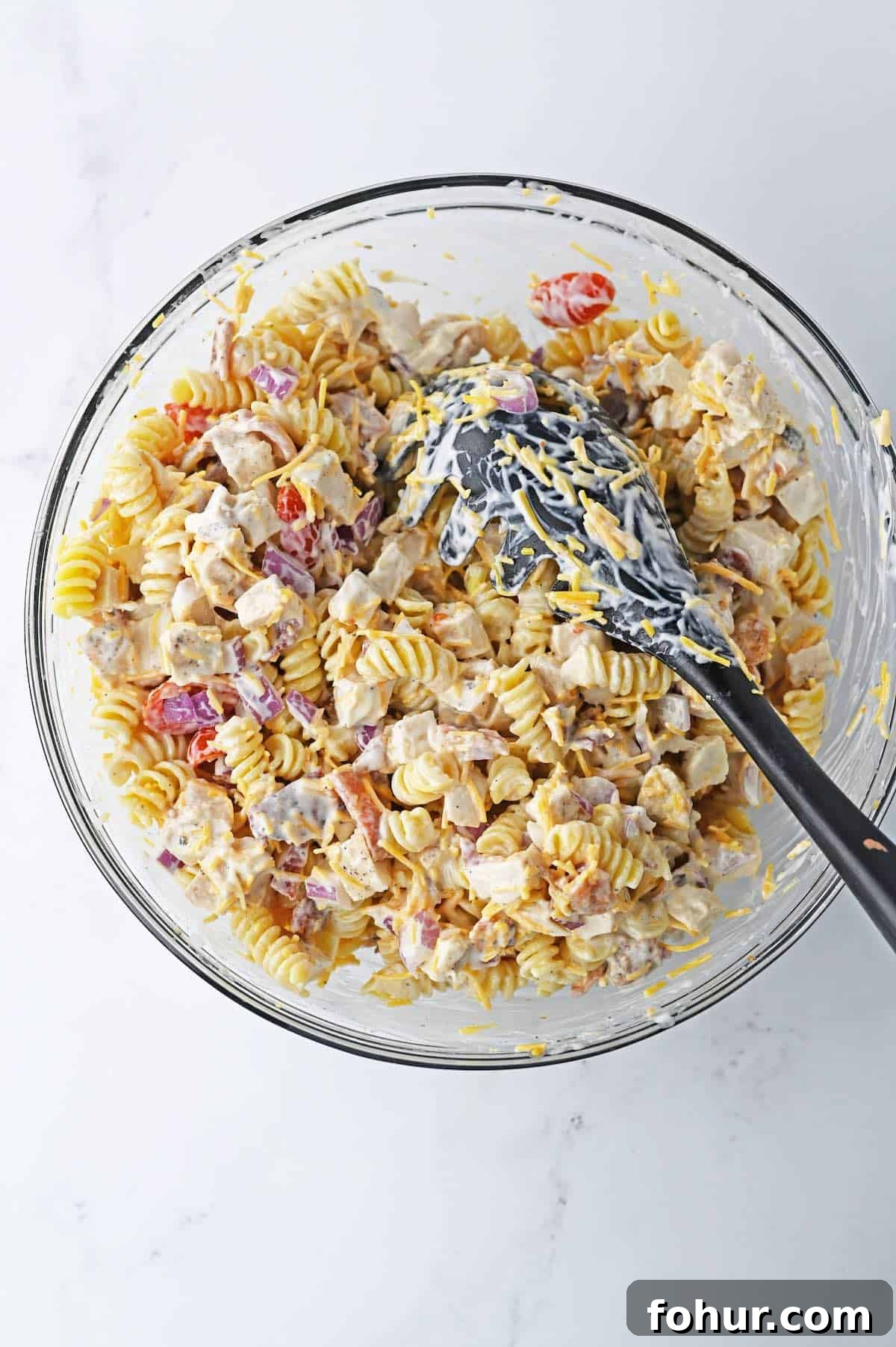 A bowl of cooked rotini noodles tossed in ranch dressing with chunks of chicken, bacon, tomatoes, red onion and shredded cheese.