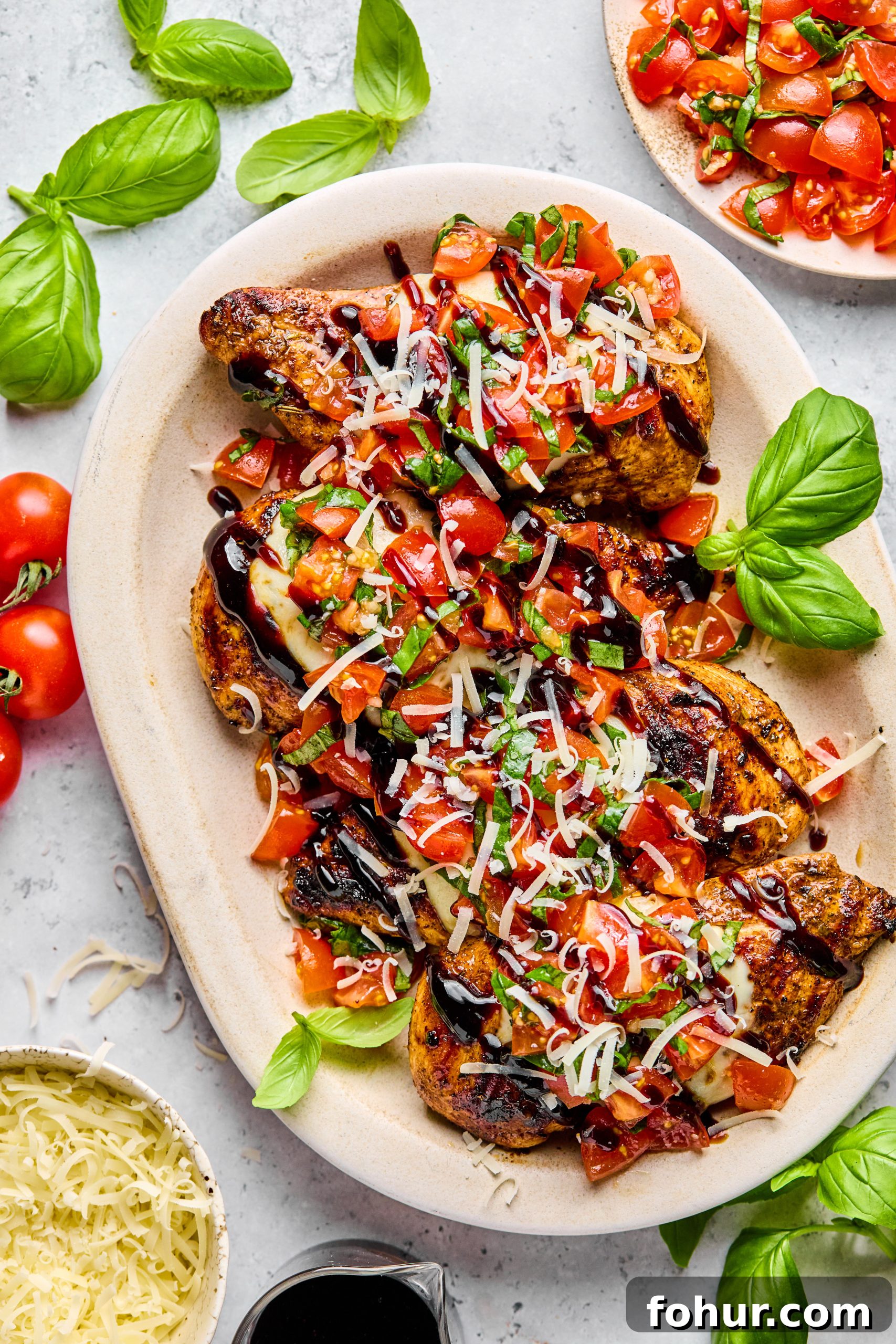 Bruschetta Chicken Recipe with Mozzarella and Balsamic Glaze 6 A serving tray filled with 4 grilled bruschetta chicken breasts sitting next to a small jar of balsamic glaze and a small bowl of shredded parmesan