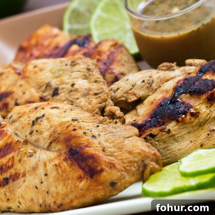 Grilled chicken on a square plate, garnished with lime slices. A small clear glass cup of Dijon basil lime dipping sauce is on the plate.