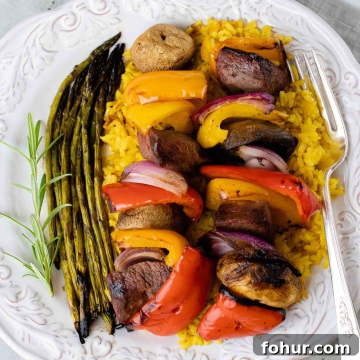 Grilled kabobs of red, yellow, and orange bell pepper pieces alternated with mushrooms, red onion pieces, and marinated beef and layered over yellow rice with grilled asparagus on a white plate with a silver fork