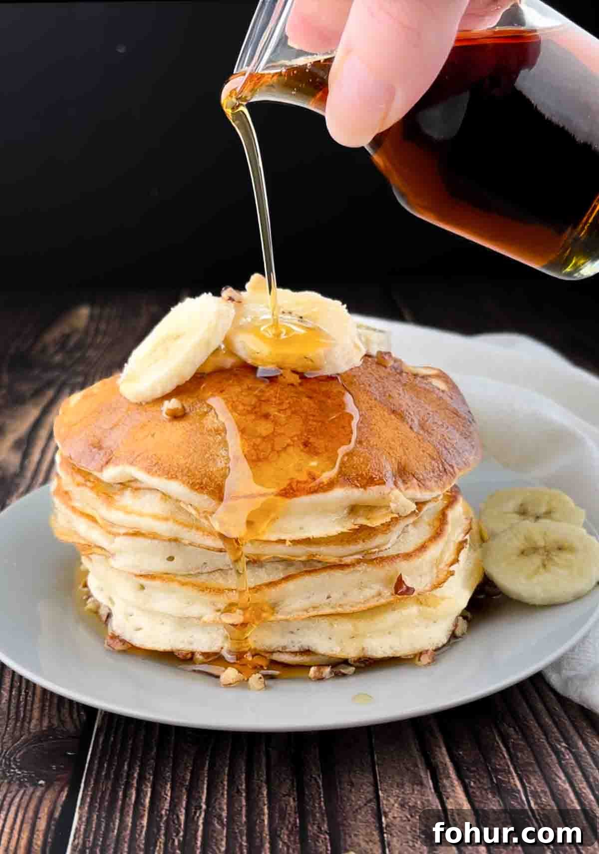 How to Make Fluffy Banana Pancakes 12 Maple syrup is drizzled over top of a stack of banana pancakes topped with sliced bananas.