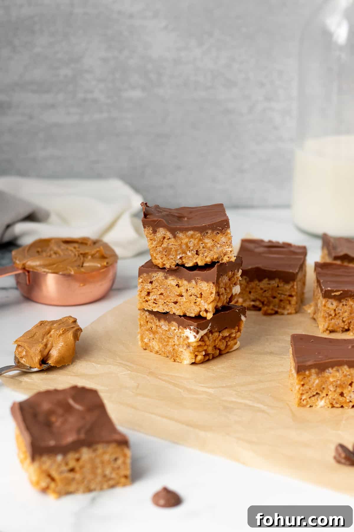 rice krispie treats on brown parchment paper with measuring cup and spoon of peanut butter and glass of milk in background