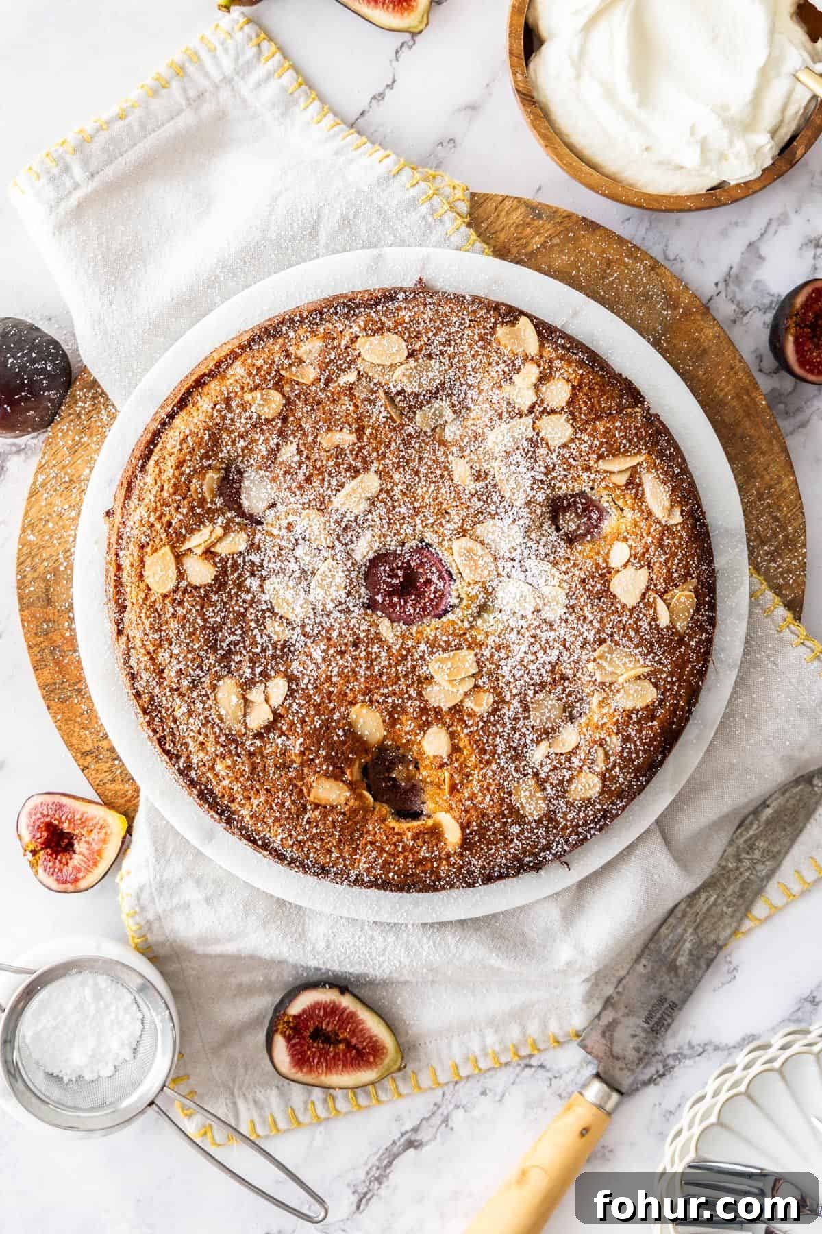 Whole Gluten-Free Fig Cake dusted with icing sugar, on a white plate, sitting on a white cloth with some figs around the edge.