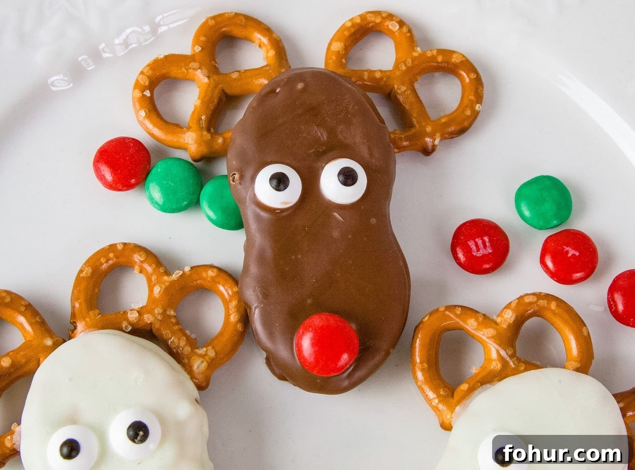 Chocolate covered Nutter Butter, surrounded by white reindeer and decorated with pretzels, M&Ms, and candy eyes. On a white plate, surrounded by red and green M&Ms
