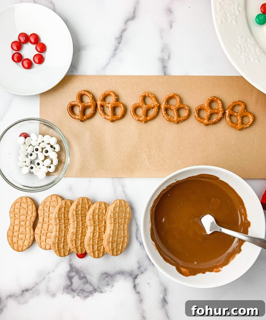 Melted chocolate in a bowl, along side Nutter Butter cookies, pretzels on brown parchment paper, a bowl of candy eyes, and red M&Ms