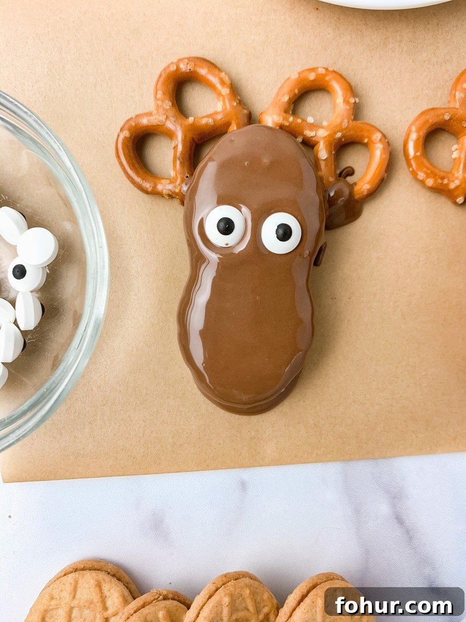 A chocolate dipped Nutter Butter Reindeer sitting on two pretzel antlers on brown parchment paper, two candy eyes added to make a face.