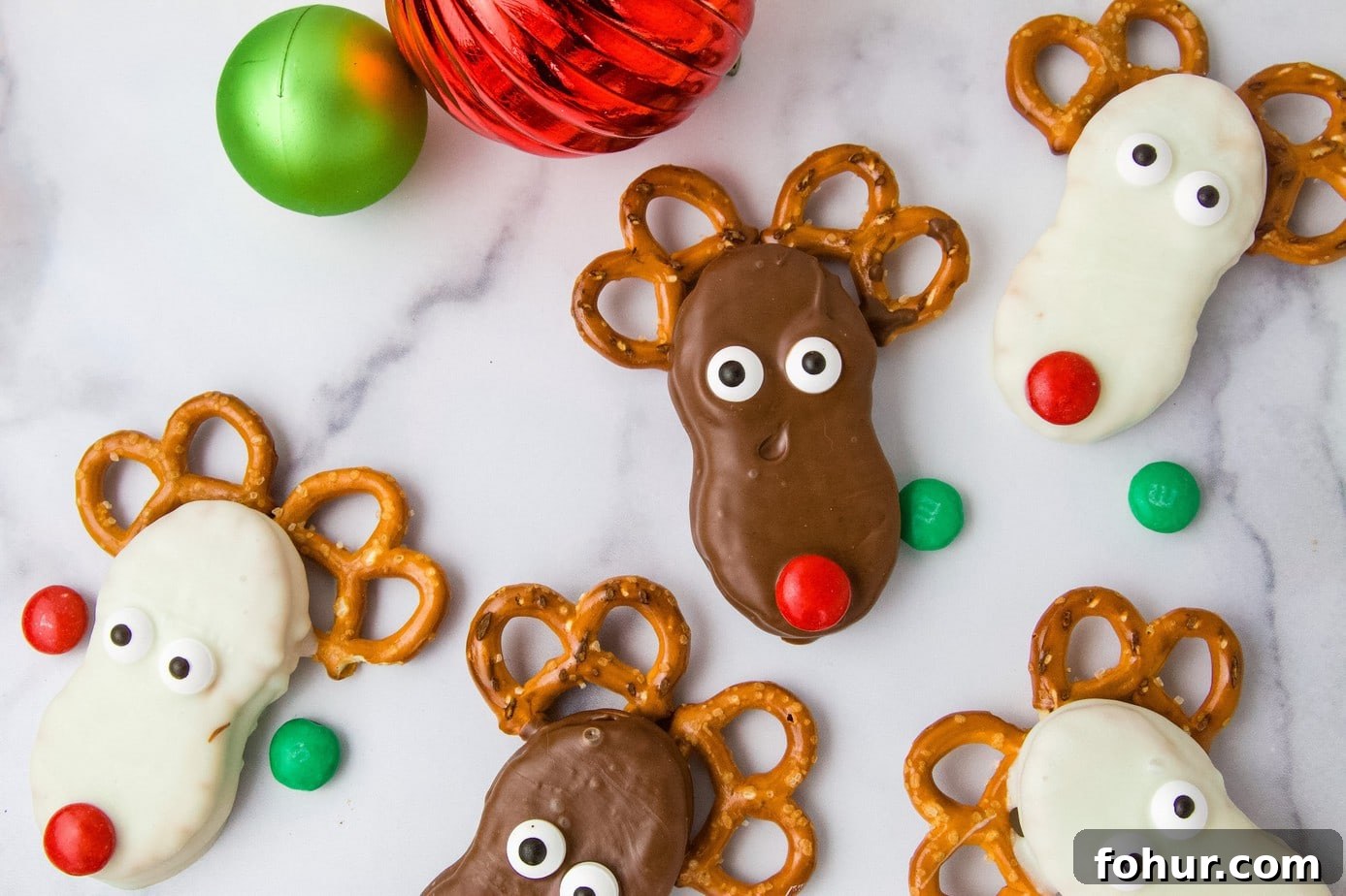 Finished white candy and milk chocolate coated reindeer, decorated with M&Ms, candy eyes, and pretzels. Lying on a marble surface with red and green M&Ms and Christmas ornaments.