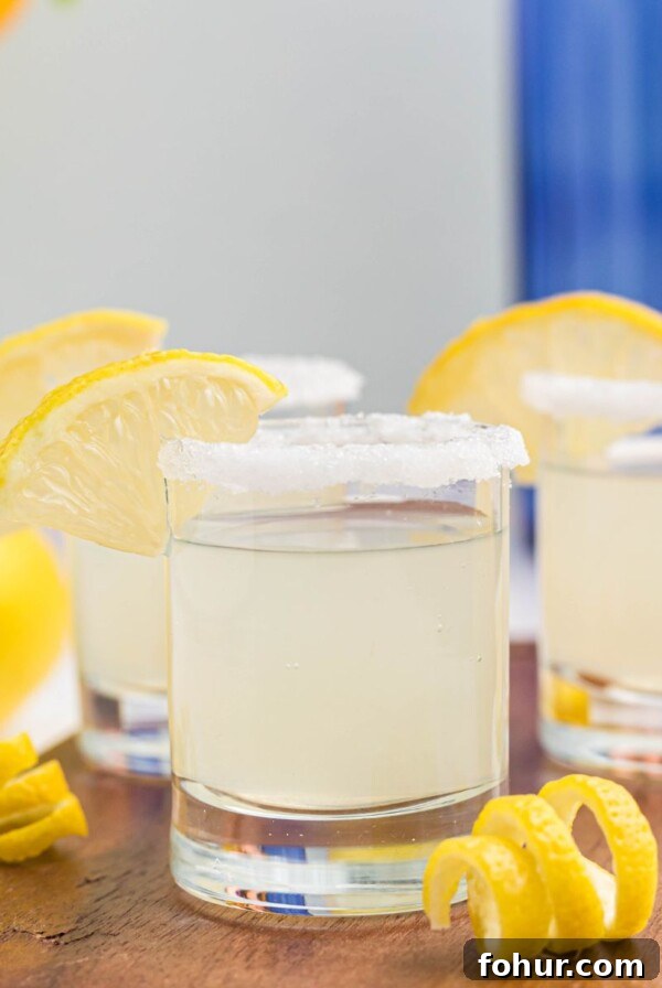 Sugar rimmed shot glass with lemon wedge for garnish, and lemon curls in front of glasses, after being filled with lemon drop shots.