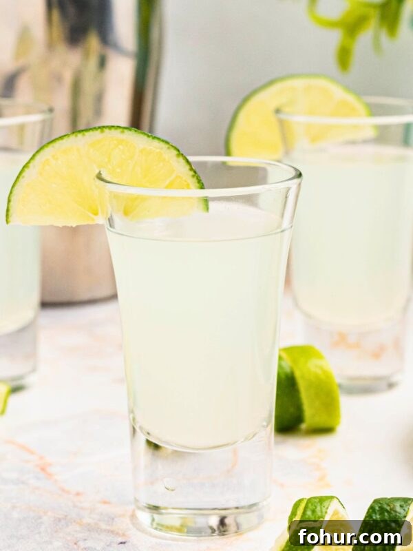 Cold Kamikaze shots in shot glasses with lime wedges and lime curls on a marble table in front of a cocktail shaker.