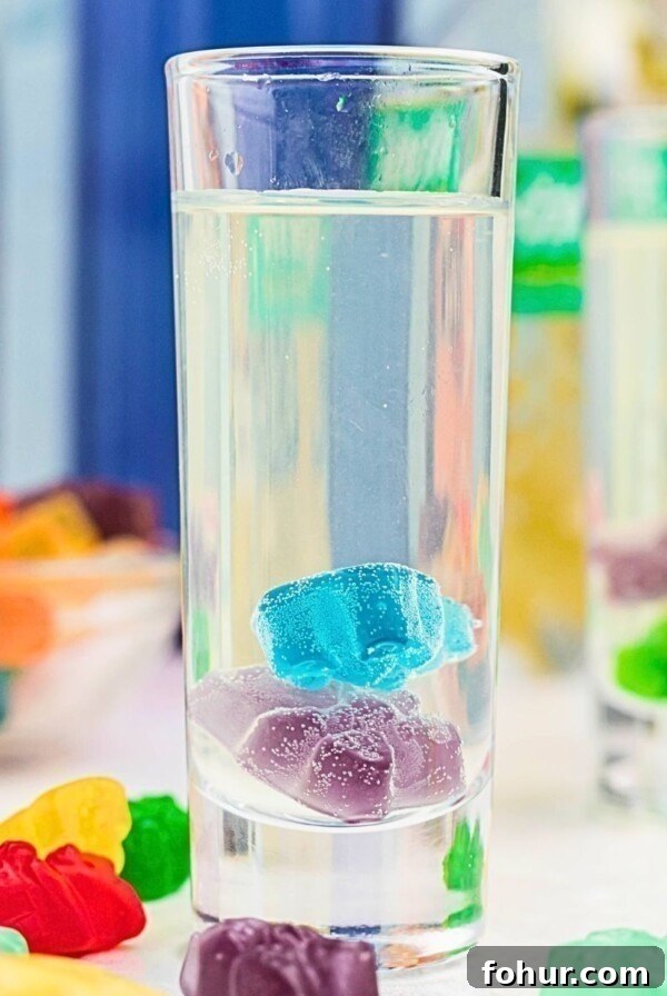 Purple and blue gummy bear in a clear shot glass with a vodka mixture poured over them, and with more gummy bears scattered on the table.
