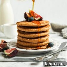 Hand pouring maple syrup from a small container over a stack of healthy oatmeal pancakes.