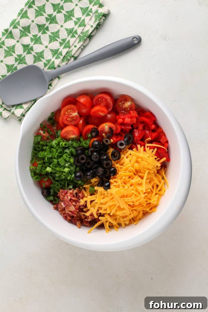 Creamy Bacon Ranch Pasta Salad with Peas and Cheddar 8 Tomatoes, cheese, bacon, and veggies being added to a white mixing bowl for pasta salad.