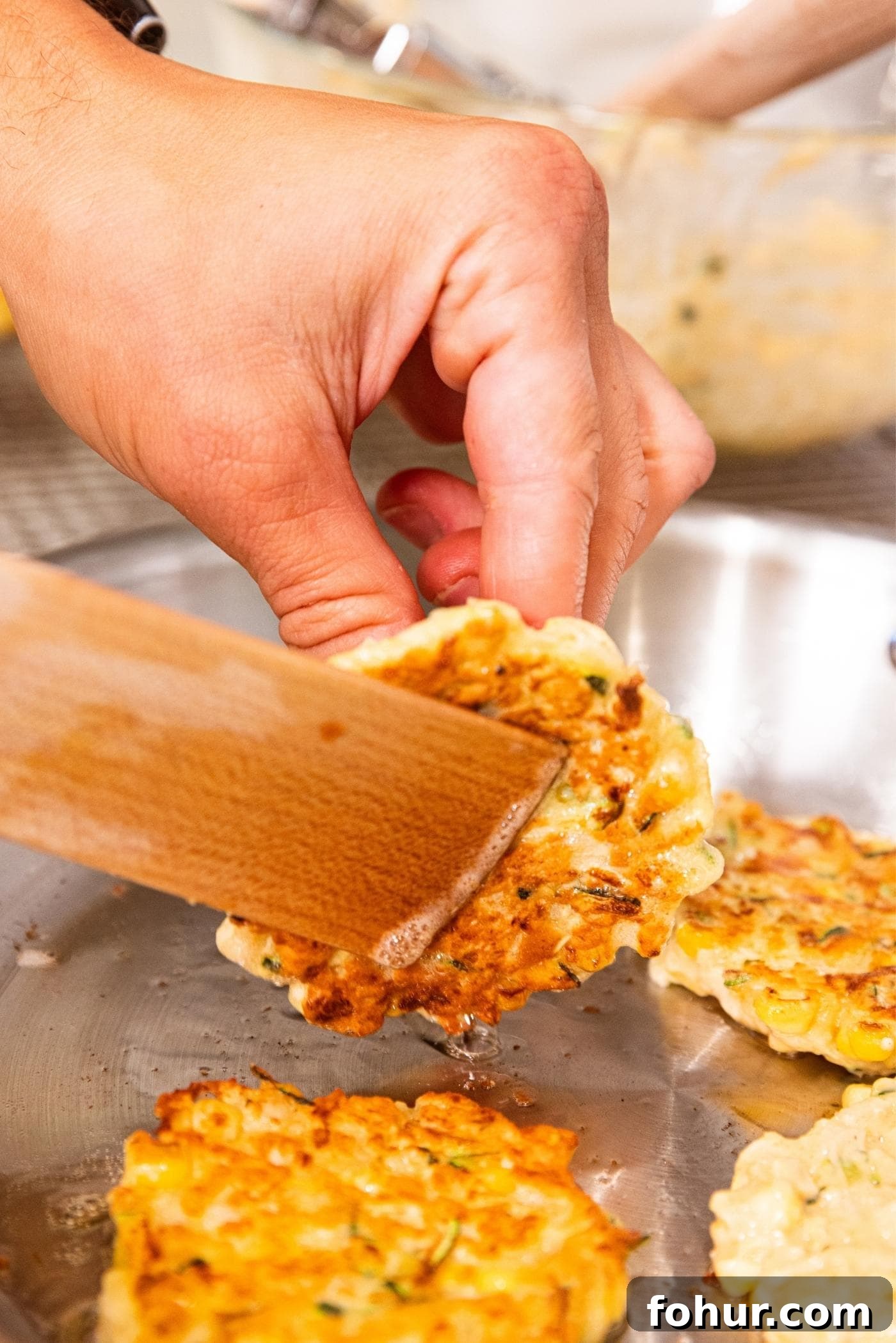 Crispy Zucchini and Sweet Corn Fritters 11 Hand using wooden spatula to flip corn fritter in metal skillet on stove.