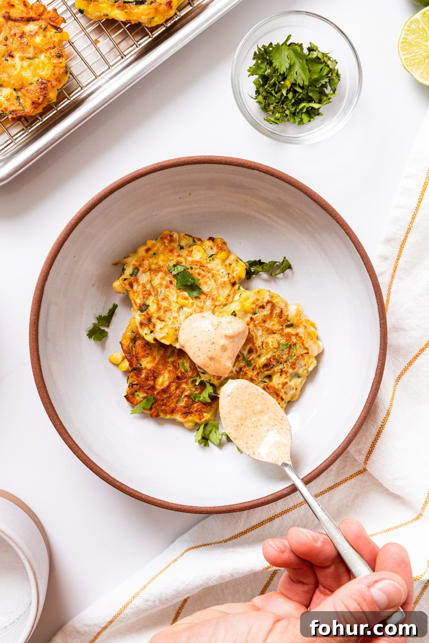 Crispy Zucchini and Sweet Corn Fritters 12 White bowl with three corn fritters topped with cilantro and spoonful of chipotle mayonnaise sauce all on white countertop.