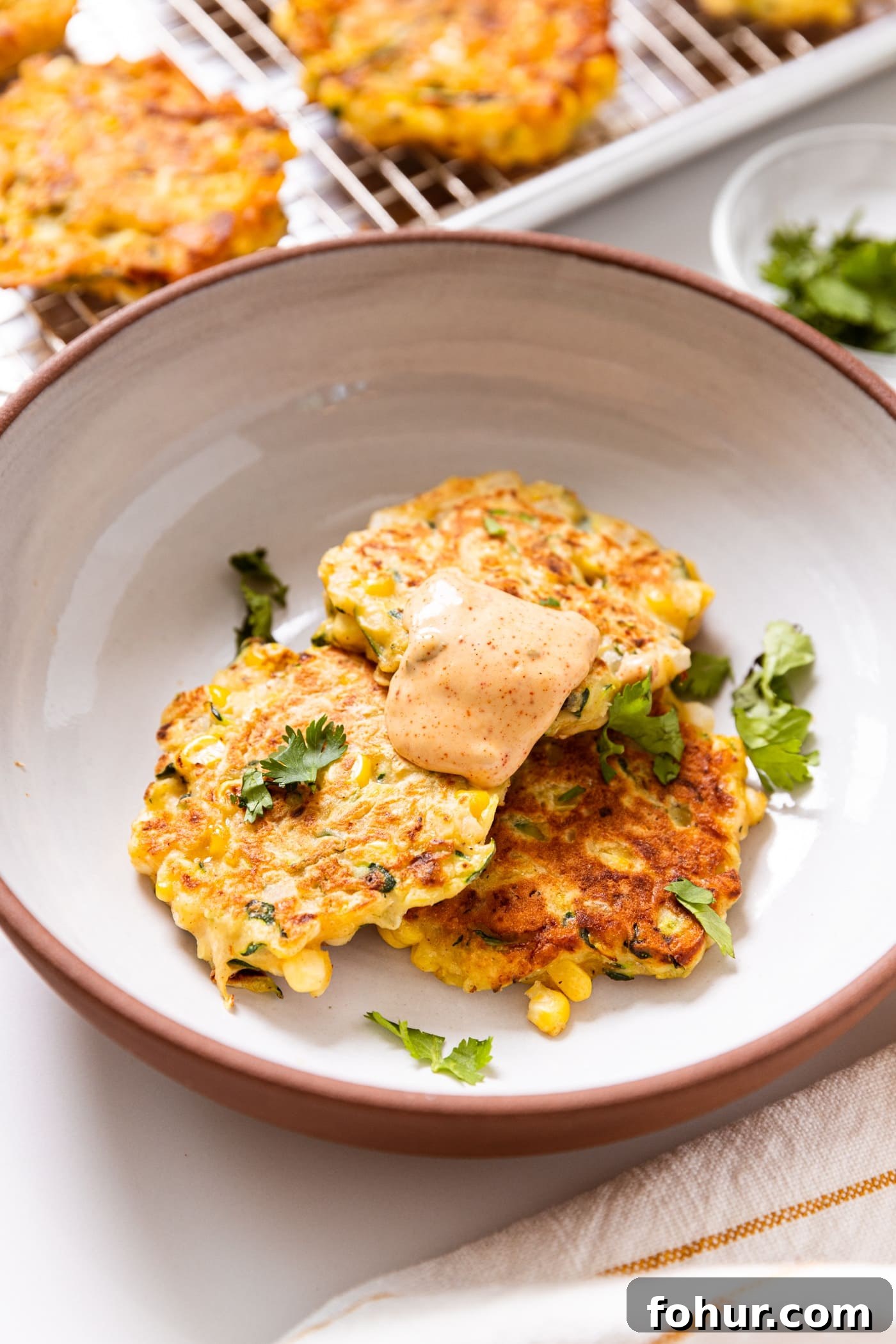 Crispy Zucchini and Sweet Corn Fritters 13 White bowl filled with three fried zucchini and corn fritters topped with chipotle mayonnaise sauce with cilantro sprinkled on top with more fritters cooling in background.