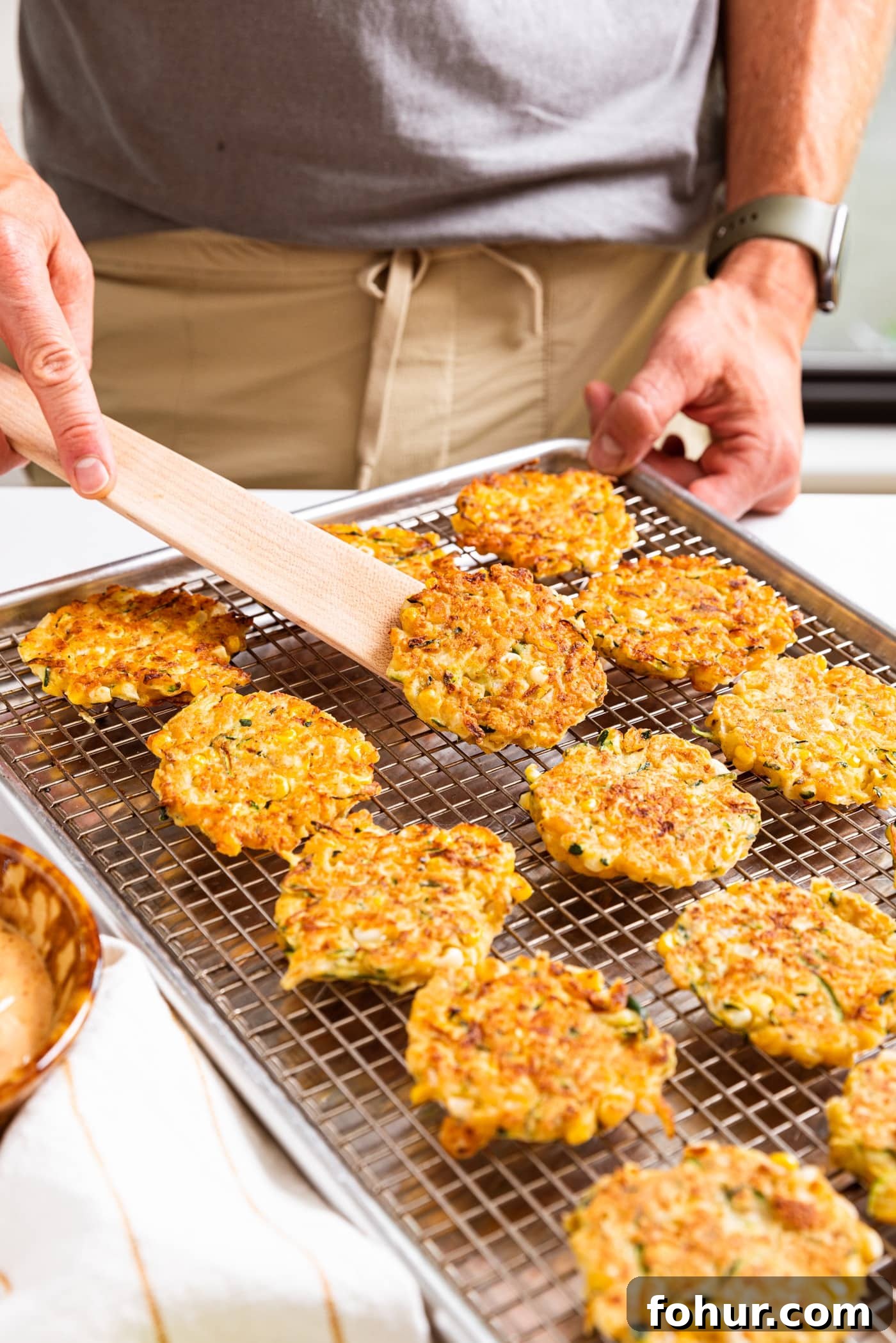 Crispy Zucchini and Sweet Corn Fritters 3 Hand holding wooden spatula scooping up one fried zucchini and corn fritter off of metal cooling rack filled with fritters all on white countertop.