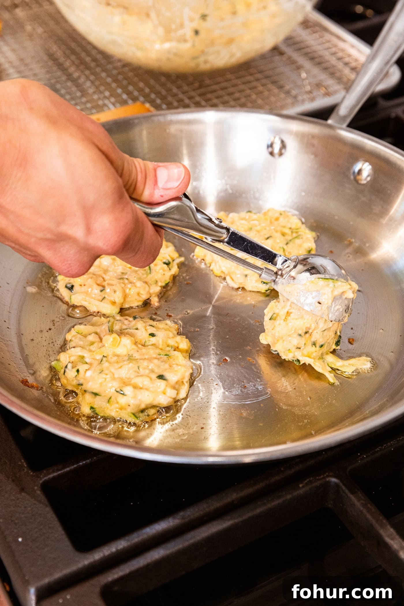 Crispy Zucchini and Sweet Corn Fritters 9 Hand dropping fritter batter from ice cream scoop onto hot metal skillet on stove.