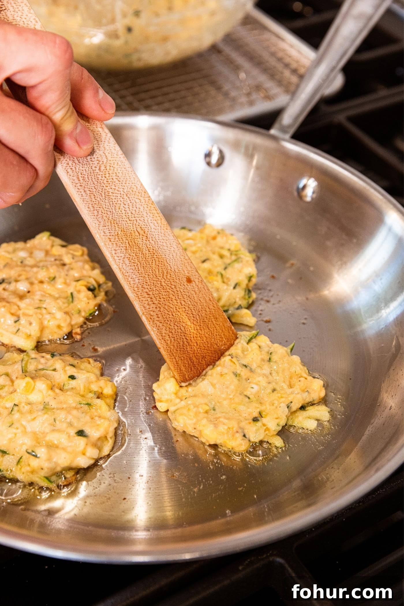 Crispy Zucchini and Sweet Corn Fritters 10 Hand pressing out fritter batter with wooden spatula on metal skillet on stove.