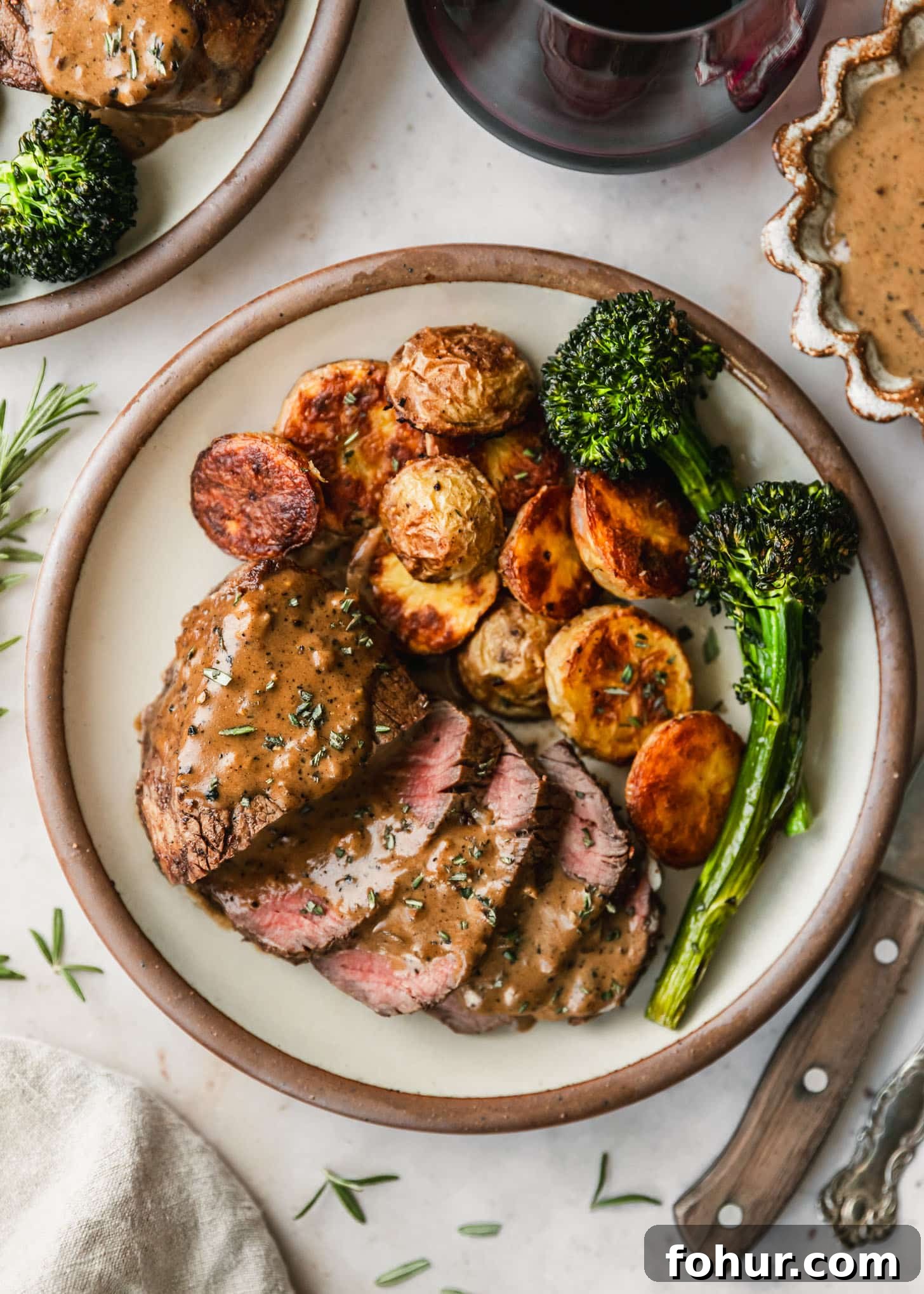 Reverse-Sear Filet Mignon Recipe with Creamy Peppercorn Sauce 7 Reverse sear steak, potatoes, and broccolini on a white plate next to red wine, a brown bowl of peppercorn sauce, and a wood steak knife on a marble counter.