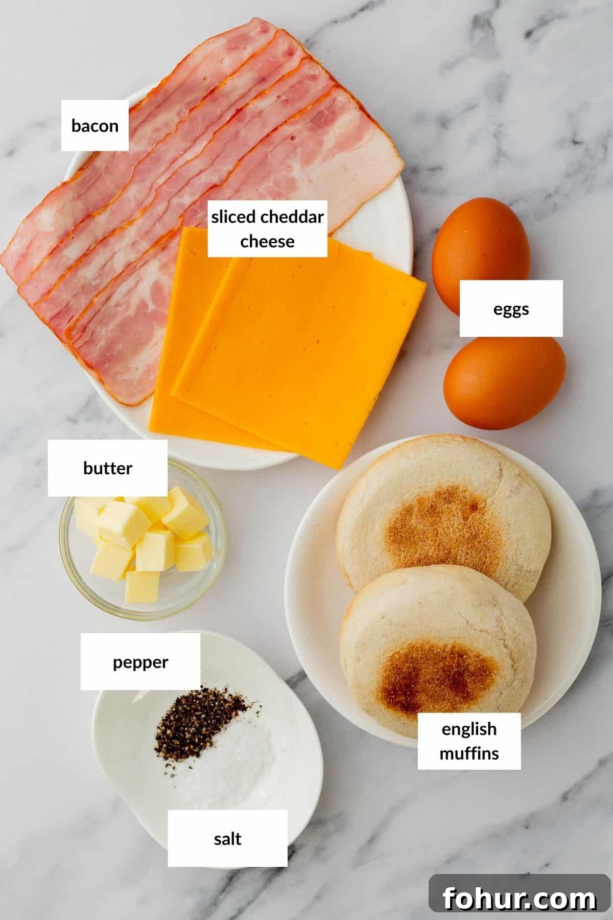 Crispy English Muffin Breakfast Sandwich with Egg and Cheese 3 A plate with bacon, sliced cheddar cheese, and two eggs, plus bowls of butter, salt, and pepper, and two English muffins on a marble surface.