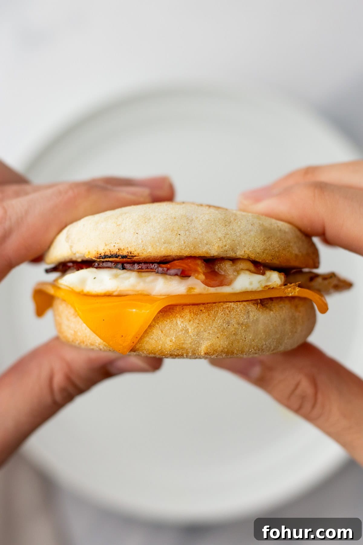 Crispy English Muffin Breakfast Sandwich with Egg and Cheese 4 A person holds a breakfast sandwich with egg, cheddar cheese, and bacon between two halves of an English muffin.