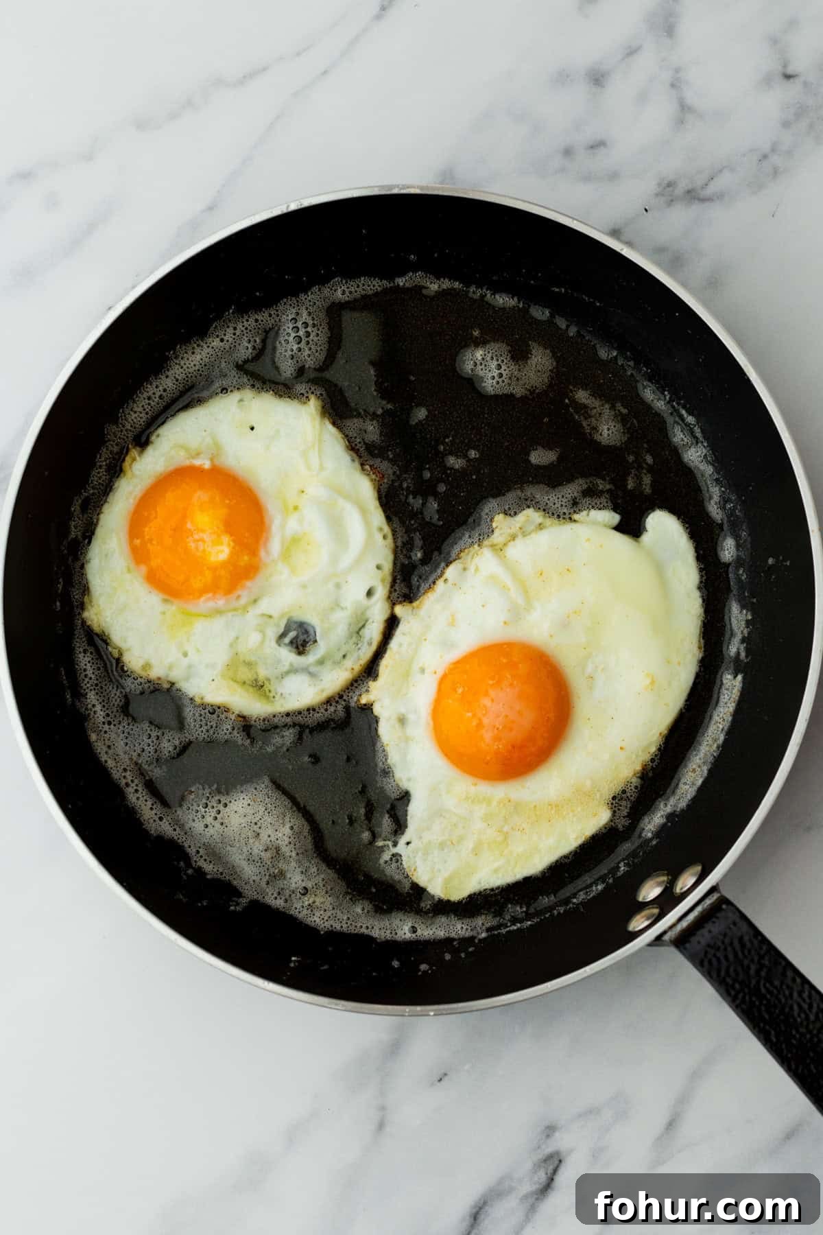 Crispy English Muffin Breakfast Sandwich with Egg and Cheese 5 Two sunny-side up eggs frying in a black skillet on a white marble surface.