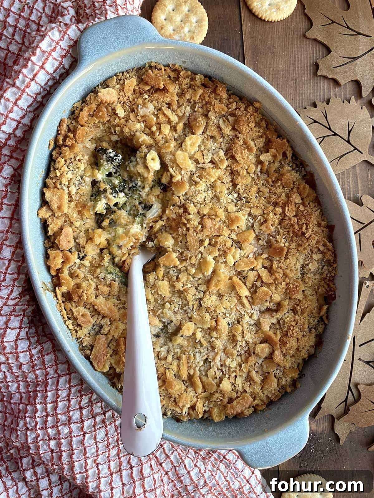 Creamy Southern Broccoli Casserole Recipe 8 A blue casserole dish filled with baked broccoli casserole topped with ritz crackers with a white serving spoon scooping some out.