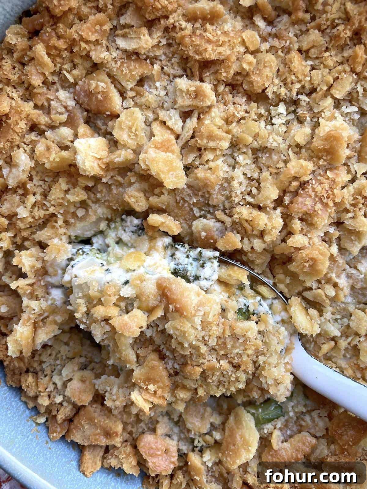 Creamy Southern Broccoli Casserole Recipe 3 A blue casserole dish filled with baked broccoli casserole topped with ritz crackers with a white serving spoon scooping some out.
