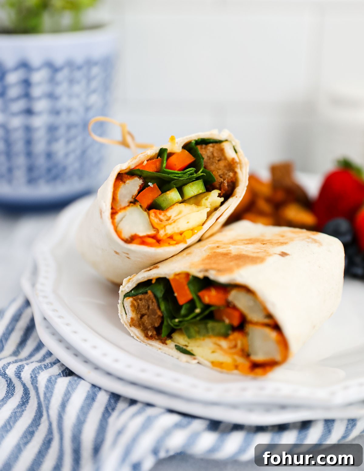 Vegan Breakfast Burrito Recipe with Tofu, Beans and Avocado 2 A plant based breakfast burrito filled with plant based egg and sausage, fresh vegetables, and breakfast potatoes. It is cut in half and on a plate with berries