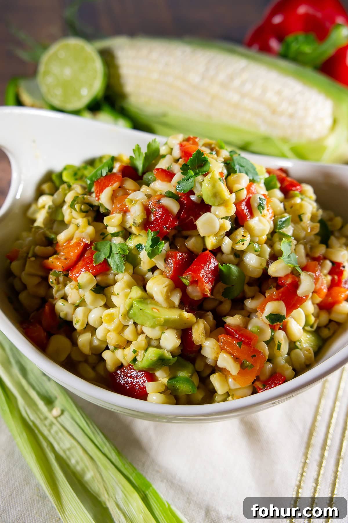 a white bowl with corn avocado salad in it with fresh corn and peppers behind it