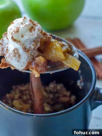spoon with apple crisp being held up above a black mug apple crisp with 2 green apples and cinnamon sticks in the background