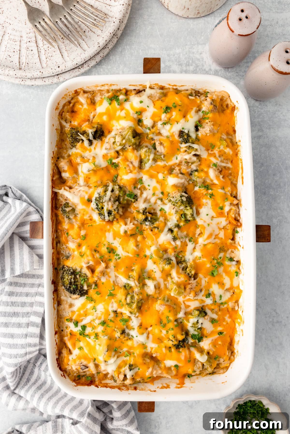 Creamy Cheesy Chicken Broccoli and Rice Casserole 5 A Cheesy Chicken Broccoli and Rice Casserole topped with melted cheddar and mozzarella, featuring visible broccoli pieces in a white dish on a light surface beside a striped towel, plate, forks, and salt and pepper shakers.