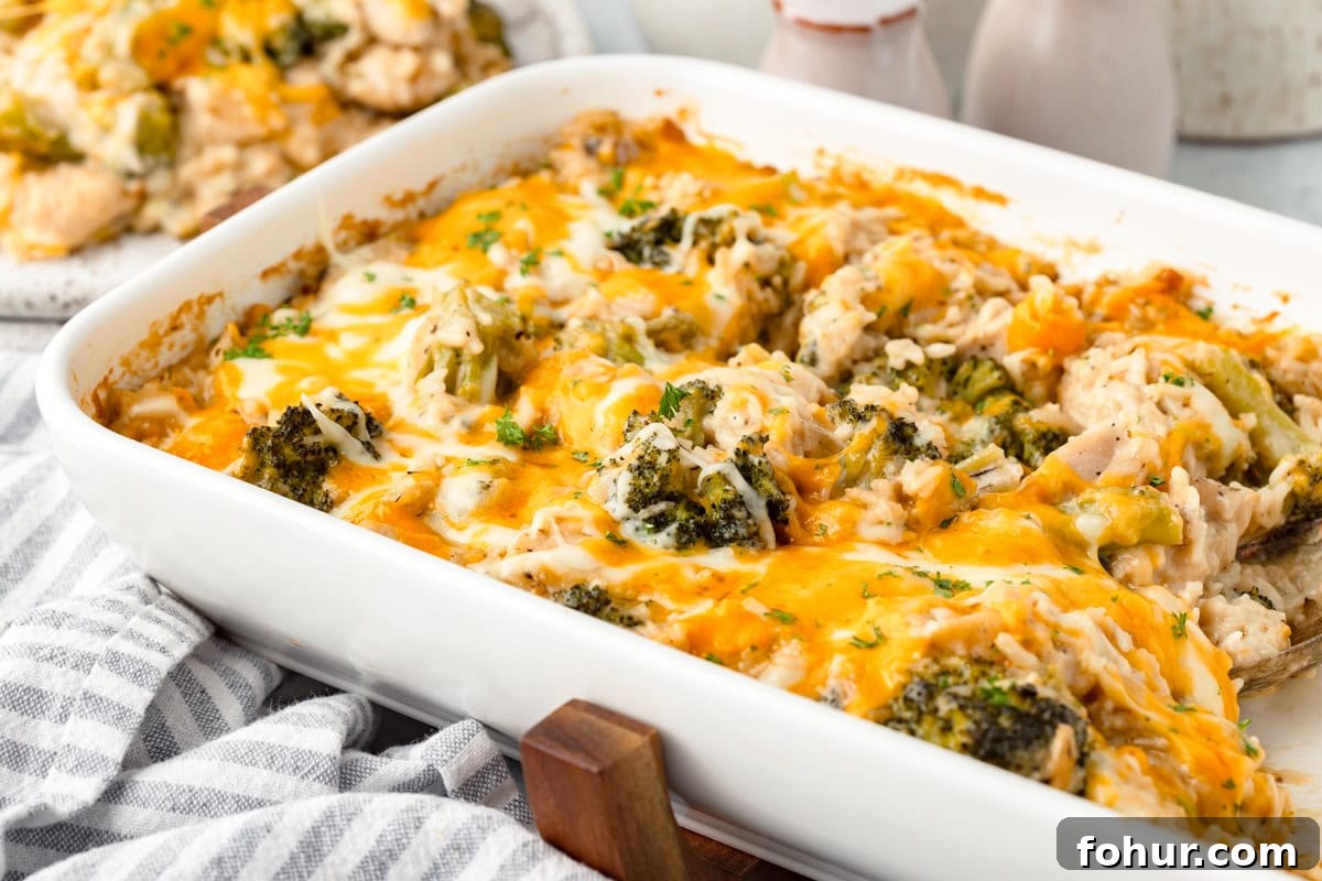 Creamy Cheesy Chicken Broccoli and Rice Casserole 6 A white baking dish filled with Cheesy Chicken Broccoli and Rice Casserole, topped with melted cheddar, sits on a striped towel. Broccoli florets and shredded chicken peek out from under the golden cheese.