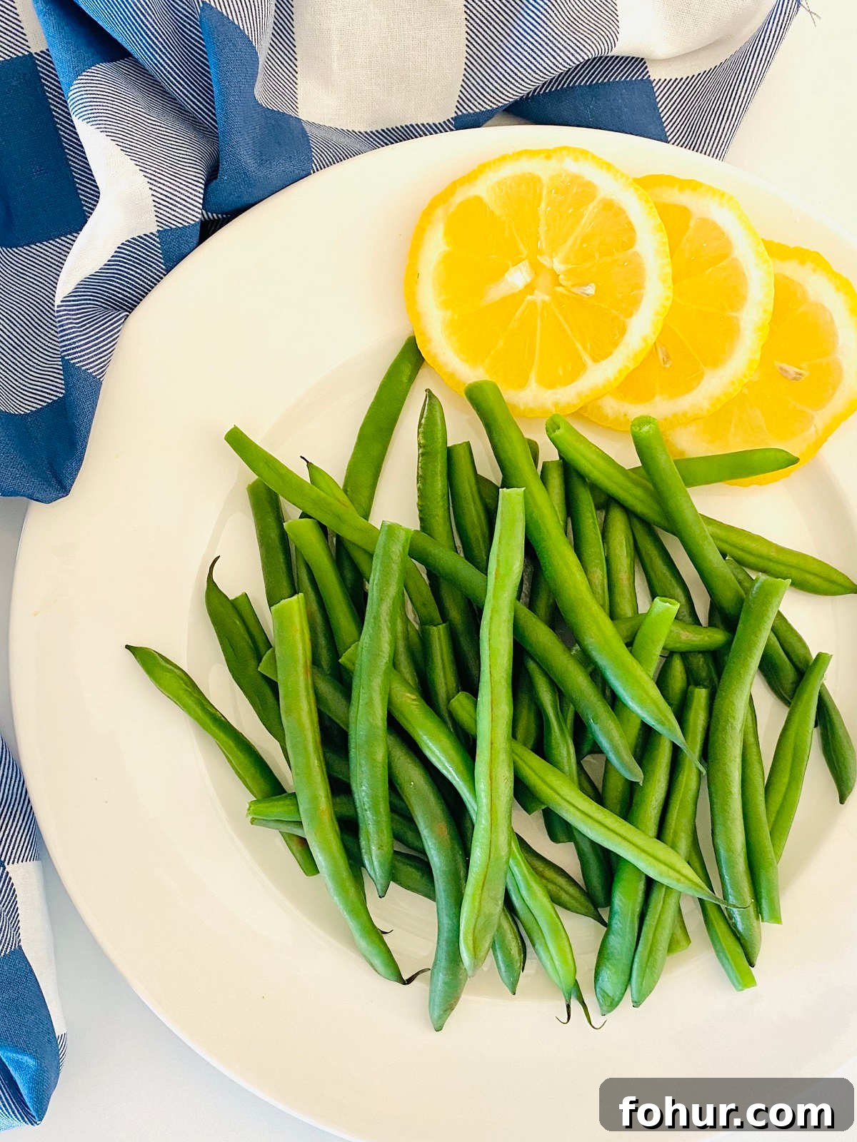 Green Bean Cooking Times: Boil, Air Fryer, Oven, Fresh & Frozen 2 green beans on a plate next to lemon slices and a blue checkered cloth napkin