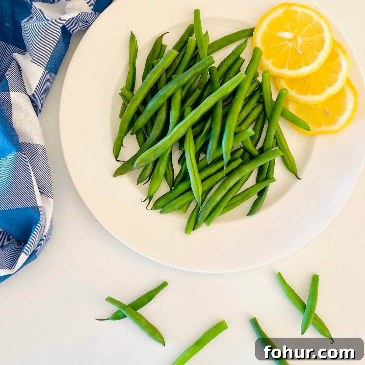 Green Bean Cooking Times: Boil, Air Fryer, Oven, Fresh & Frozen 3 green beans on a plate next to lemon slices and a blue checkered cloth napkin