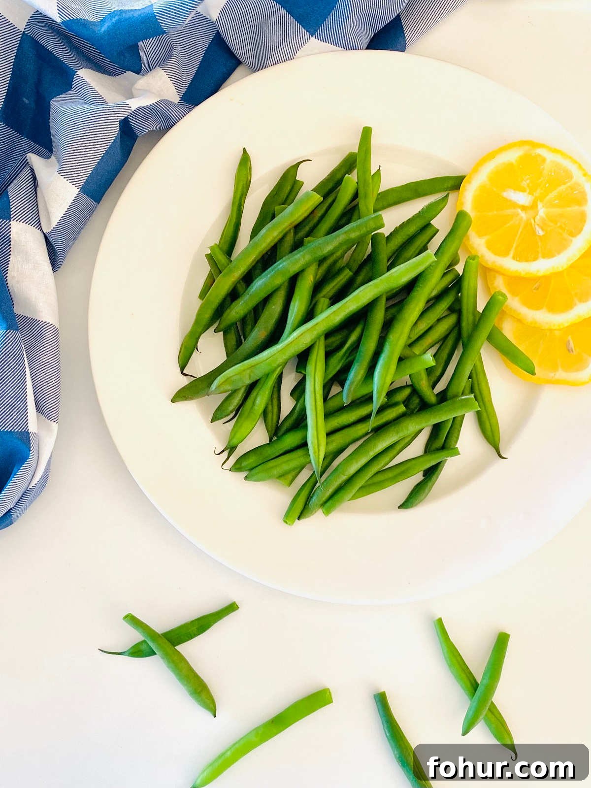 Green Bean Cooking Times: Boil, Air Fryer, Oven, Fresh & Frozen 4 green beans on a plate next to lemon slices and a blue checkered cloth napkin