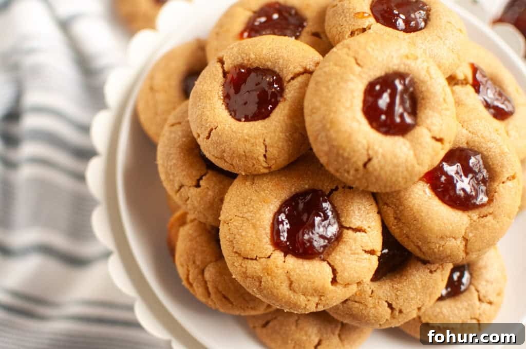 19 Cookie Recipes Even Dessert Skeptics Can't Resist 4 A plate stacked with Peanut butter jelly thumbprint cookies filled with red jam, placed on a white stand next to a striped cloth.